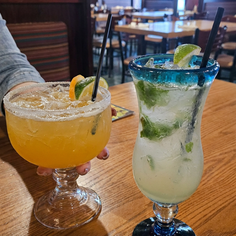 Margarita and mojito garnished with lime sit on a wooden restaurant table, with diners and seating in the background.