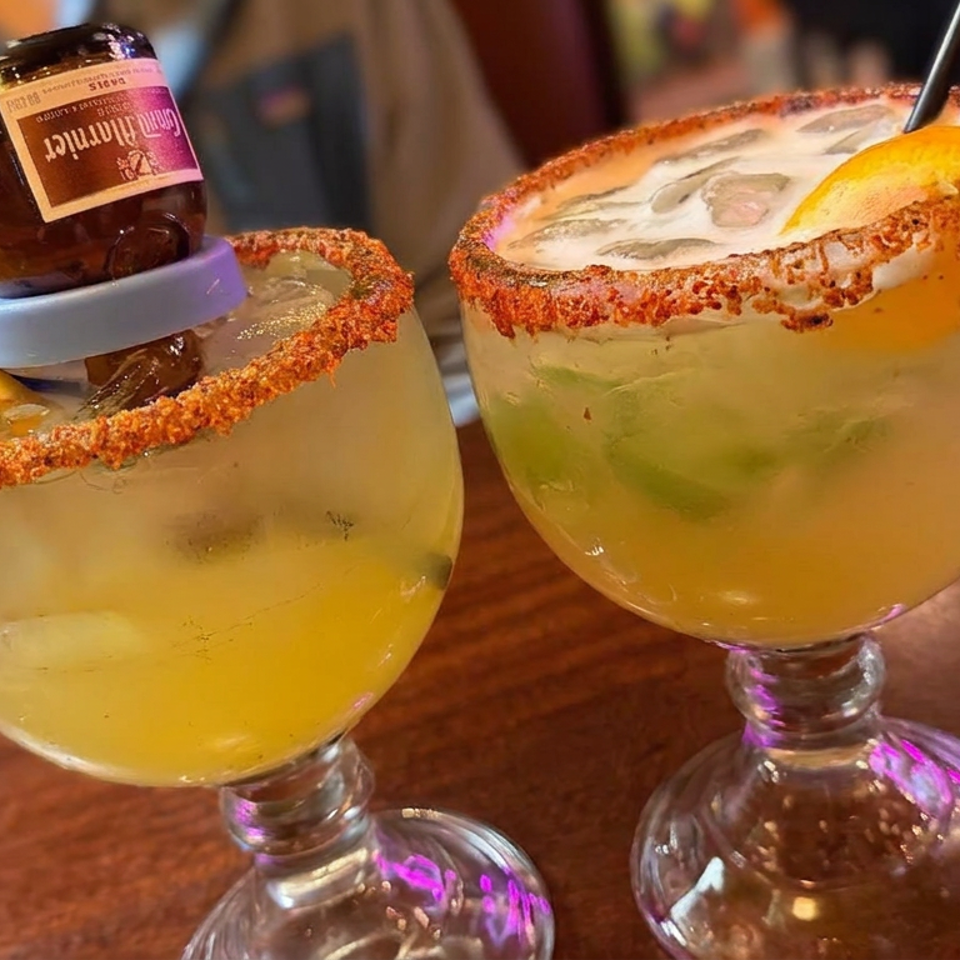Two citrus margaritas with chili-salt rims, orange wedges, lime, and an inverted mini beer bottle on a restaurant table.