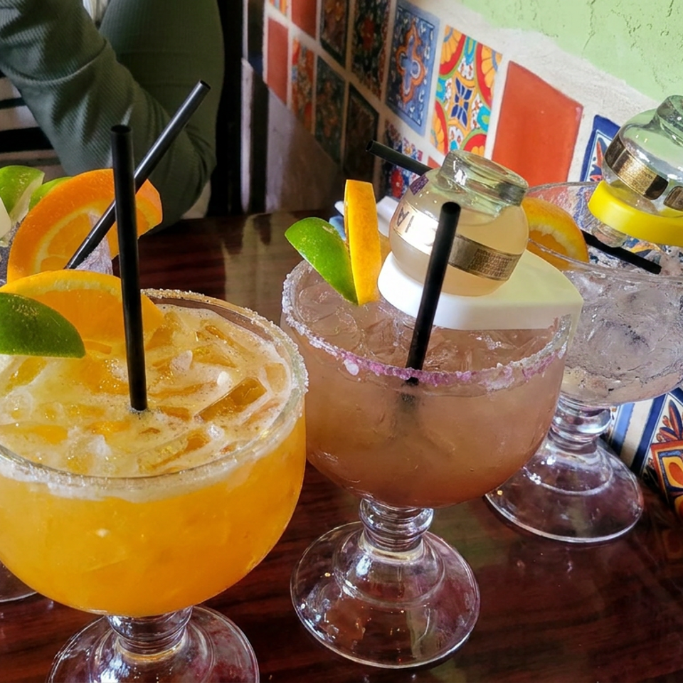 Four colorful cocktails with citrus garnishes and black straws sit on a wooden table beside vibrant patterned tiles.