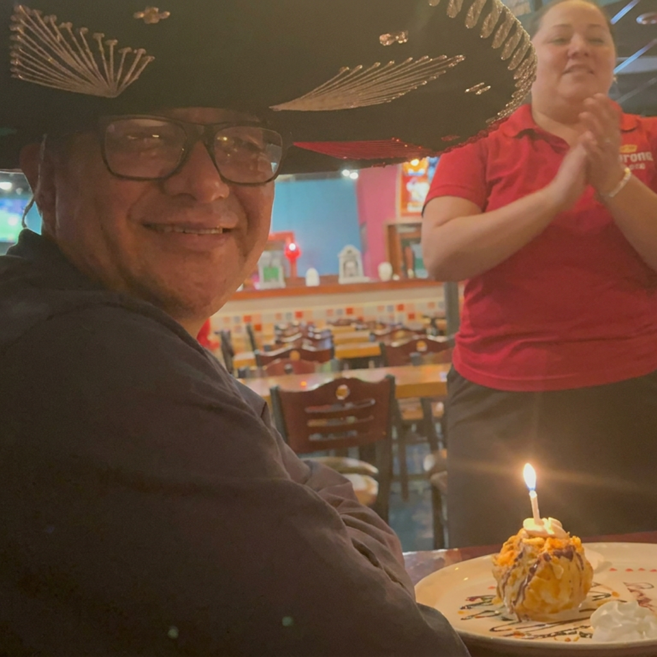 Diner wearing a sombrero smiles beside a lit birthday dessert while a restaurant server claps behind him.