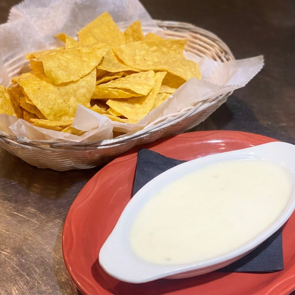 Basket of tortilla chips beside a white dish of creamy queso on a red plate atop a restaurant table.